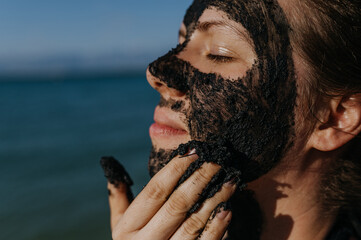 Woman applies healing mud to her face and body. Natural healing mud in Croatia on the beach. - obrazy, fototapety, plakaty