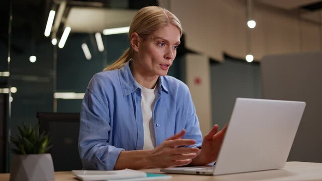Concentrated female dressed in informal clothes sitting at office desk and typing on modern laptop. Blond woman getting upset while working at computer because of problem with internet connection.