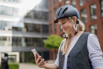 Beautiful middle-aged woman commuting through the city by bike, holding smartphone. Female city...