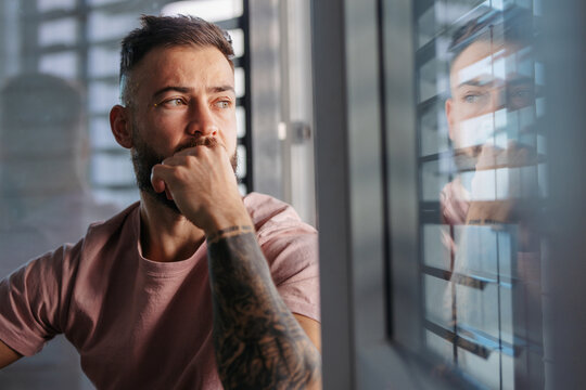 Calm man in pink t-shirt with tattoo on arms looking through window.