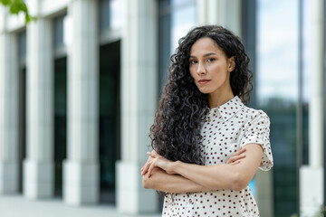 Professional young businesswoman with curly hair standing confidently outside an office, arms...