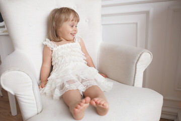 A little girl in a white lace dress is sitting on a white chair.
