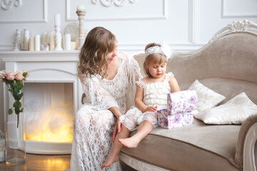 A mother and a daughter in lace dresses are sitting on a sofa in the living room. White interior. 