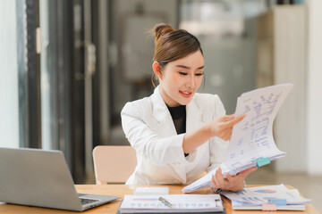 A portrait of a beautiful and confident Asian businesswoman in a pink business suit sits at her desk in the office with her arms crossed, smiling at the camera.