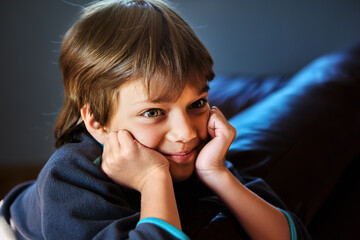 Young boy with brown hair and green eyes is sitting on a couch. He has his hands propped under his...