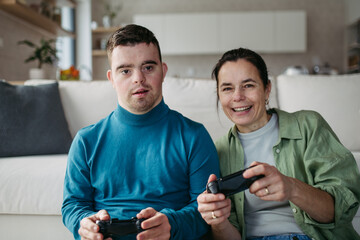 Young man with Down syndrome with his mother playing video game, holding game controllers. Concept...
