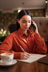 Beautiful woman in cafe, drinking coffee, writing in notebook, checking documents on lunch break, call for client on mobile phone.