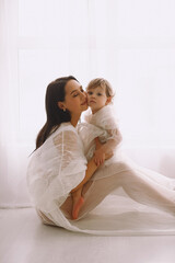 Studio portrait of happy mother sitting near window and hugging her baby girl. Mother and daughter in white dresses.