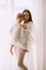 Happy mother in white dress standing near window and holding her baby daughter in her arms