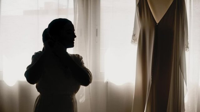 Silhouette of bride looking at her dress as light enters through window. Slow motion