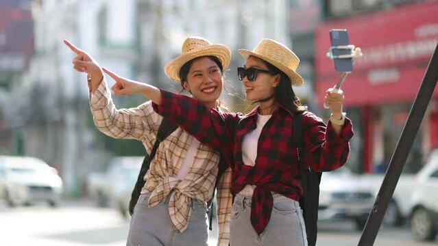 Two female Asian tourist friends are taking pictures and selfies together on a sidewalk along the side of a road in Thailand.