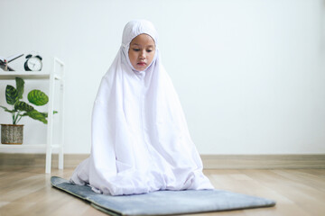 Asian Muslim little girl doing one of movement gesture in salat procedure. The worshiper proceeds to sit and recite prayers - obrazy, fototapety, plakaty