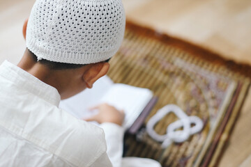 Rear view of muslim kid wear skullcap, reading Quran on prayer mat at the mosque