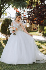 Fototapeta premium Wedding photo. A brunette bride in a lacy voluminous dress, holding a bouquet and walking in the garden, smiling sincerely. Gorgeous hair and makeup. Fashion and style. Celebration.
