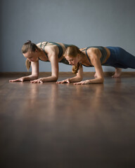 Fototapeta na wymiar Two girls doing yoga, fitness in gym. Elbow plank exercise. ardha pincha mayurasana, dolphin pose. Training back, abdominal muscles, core muscles. Wooden reflective floor, gray background. Side view.
