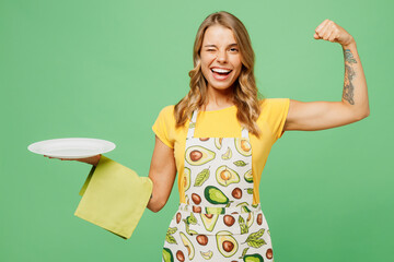 Young strong cool happy housewife housekeeper chef cook baker woman wears apron yellow t-shirt hold in hand empty plate towel show hand biceps isolated on plain green background. Cooking food concept. - obrazy, fototapety, plakaty
