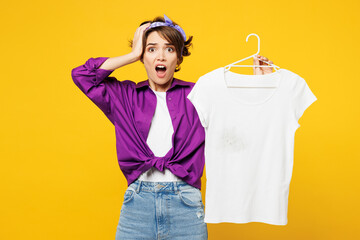 Young shocked scared astonished sad woman wears purple shirt do housework tidy up hold t-shirt on...