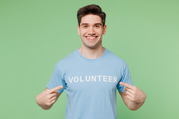 Young smiling happy man he wears blue t-shirt white title volunteer point index finger on himself isolated on plain pastel green background. Voluntary free work assistance help charity grace concept.