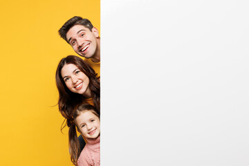 Young fun smiling parents mom dad with child kid girl 7-8 years old wear pink casual clothes hold big empty blank poster billboard for content isolated on plain yellow background. Family day concept