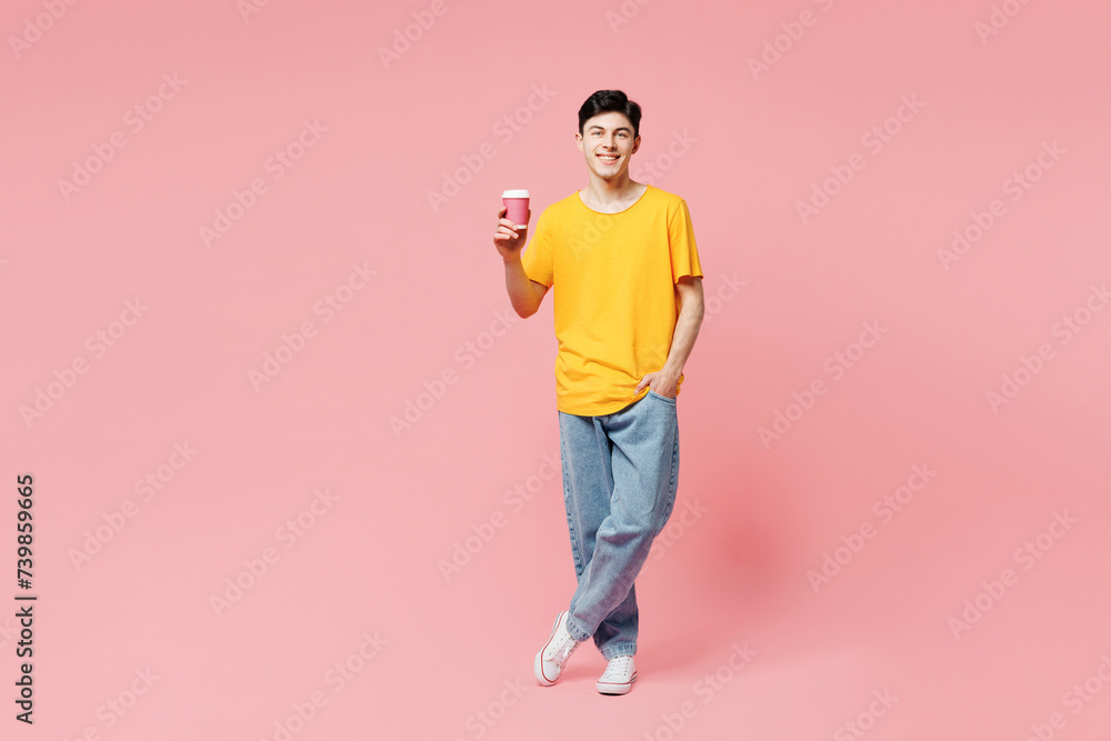 Wall mural Full body young man he wears yellow t-shirt casual clothes hold takeaway delivery craft paper brown cup coffee to go isolated on plain pastel light pink background studio portrait. Lifestyle concept.