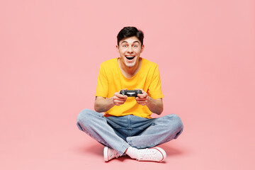Full body happy young man he wears yellow t-shirt casual clothes sits hold in hand play pc game with joystick console isolated on plain pastel light pink background studio portrait. Lifestyle concept.