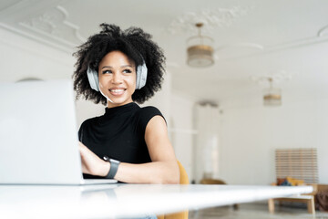 Cheerful woman with headphones working on a laptop, embodying remote work with a touch of comfort...