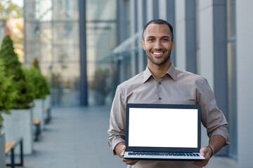 Portrait of a smiling hispanic man holding a mockup of a laptop with a white screen, standing on the street near an office building and showing the device to the camera.