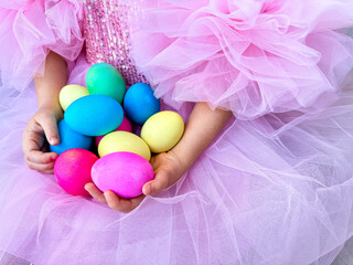 Child in pink tulle dress holds in her hands handful of bright multi colored Easter eggs, symbolizing the spring holidays and the celebration of Easter.