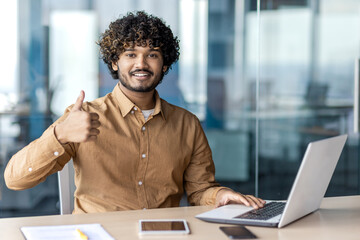 Portrait of a smiling young Indian man sitting at an office desk with a laptop, looking at the camera and showing a super success gesture with his finger.