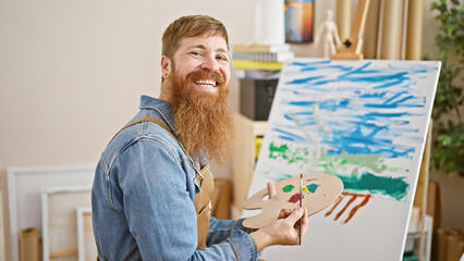 Smiling young redhead man radiating confidence, drawing away blissfully at art studio, an indoor...
