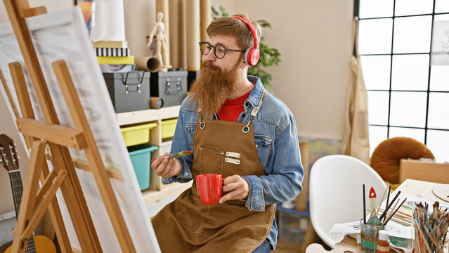 Handsome young redhead man artist, coffee in hand, drawing intently while listening to music in a bustling art studio