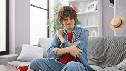 A young man winces in pain, clutching his elbow in a modern living room, signaling injury and discomfort.