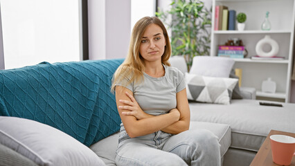 Expressive portrait of a worried young blonde woman sitting cross-armed on her living room sofa,...