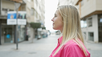 Fototapeta premium Profile of a young blonde woman in a pink shirt standing on an urban street, looking into the distance