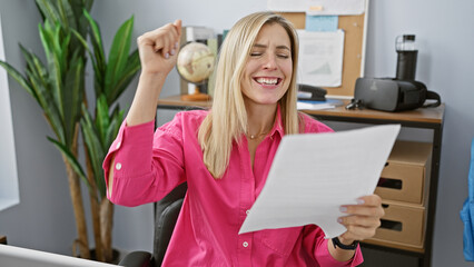 A joyful blonde woman celebrates success while reading a document in a modern office setting