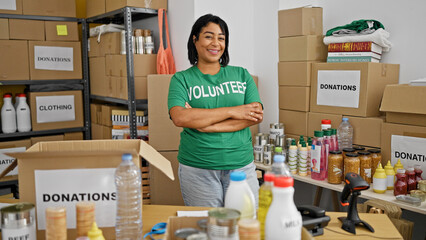 Confident hispanic woman volunteering in a donation center, surrounded by boxes and shelves of supplies.