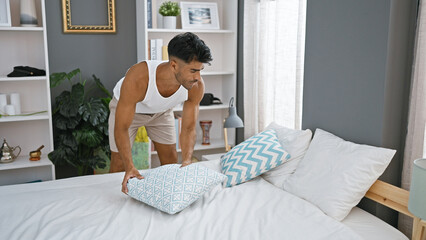 A young hispanic man arranges pillows in a modern bedroom at home, showcasing a tidy and stylish...