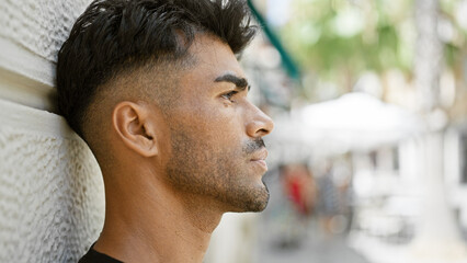 Pensive hispanic man with beard leaning on urban wall gazing down street