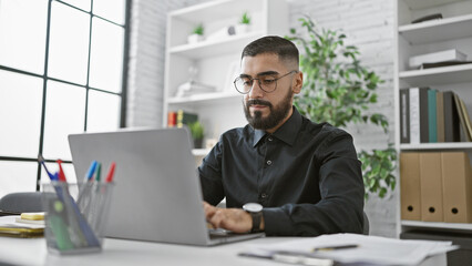 A focused bearded man working diligently on his laptop in a modern office setup.