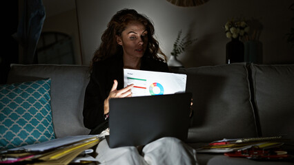A focused caucasian woman analyzes financial charts on paper while working late on her laptop in a...