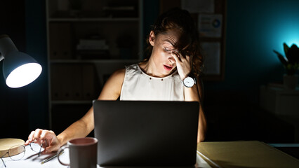 A young woman looks stressed working late in her office on a laptop, illuminating her face with a...