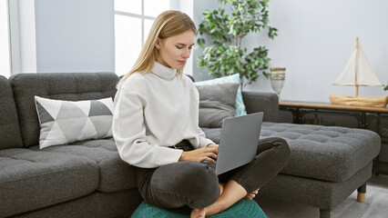 A blonde woman works on her laptop while seated comfortably in a modern living room, exuding casual elegance