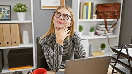 Thoughtful blonde woman with glasses and blue eyes daydreaming at office desk indoors
