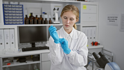 A young, focused, blonde woman wearing gloves and lab coat stands in a laboratory setting, implying...