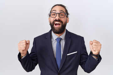 Hispanic man with beard wearing suit and tie celebrating surprised and amazed for success with arms raised and open eyes. winner concept.