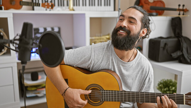 A cheerful hispanic man plays guitar in a home studio with microphones and instruments visible.