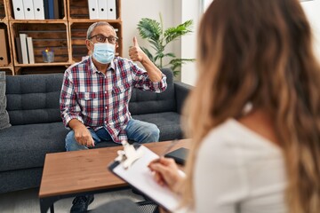 Cheerful senior man in medical mask gives cool 'ok' sign in psychology clinic. positive elder with grey hair sitting, smiling and showing confident approval gesture indoors.