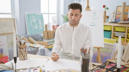 Young, handsome hispanic man ardently drawing on paper, deeply engrossed and concentrated in his...