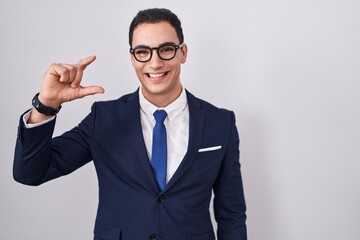 Young hispanic man wearing suit and tie smiling and confident gesturing with hand doing small size sign with fingers looking and the camera. measure concept.