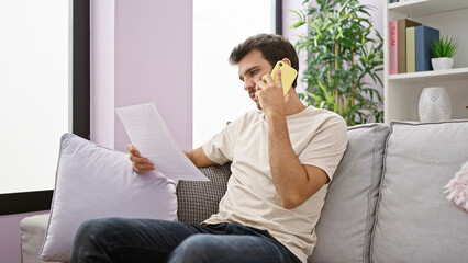 A focused young man reads a document while talking on the phone in a cozy, plant-decorated living room.